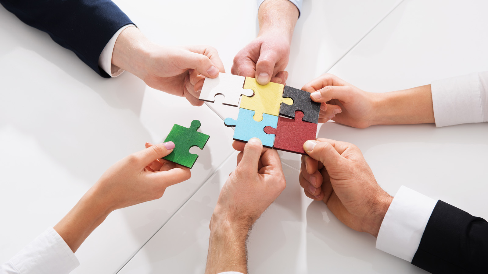 Mehrere Hände halten farbenfrohe Puzzlestücke und arbeiten zusammen daran, sie auf einer weißen Tischoberfläche zusammenzufügen.