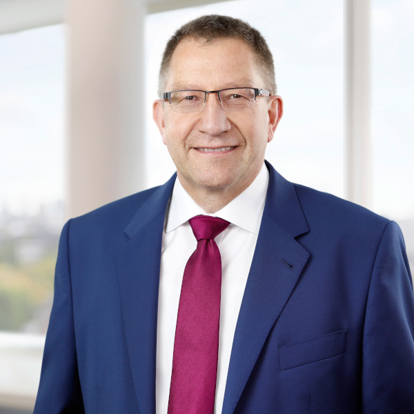 Ein Mann trägt einen blauen Anzug mit einem roten Krawatte und steht vor großen Fenstern mit Blick auf die Stadt.