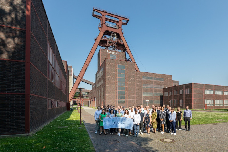 Eine Gruppe von Menschen steht vor einem alten Industriegebäude mit einem großen roten Förderturm vor einem klaren blauen Himmel.