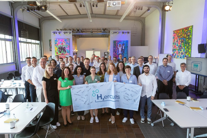 Eine Gruppe von Menschen posiert mit einem Banner für H2Hercules in einem lebendigen Veranstaltungsort mit bunten Kunstwerken.