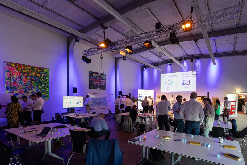 Ein Konferenzraum voller Menschen, die sich an Gesprächen beteiligen. Bildschirme zeigen Präsentationen unter lila Beleuchtung.