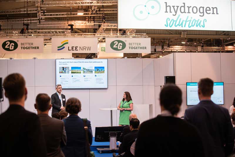 A presentation on hydrogen solutions at a trade show. A woman in a green dress stands beside a screen.