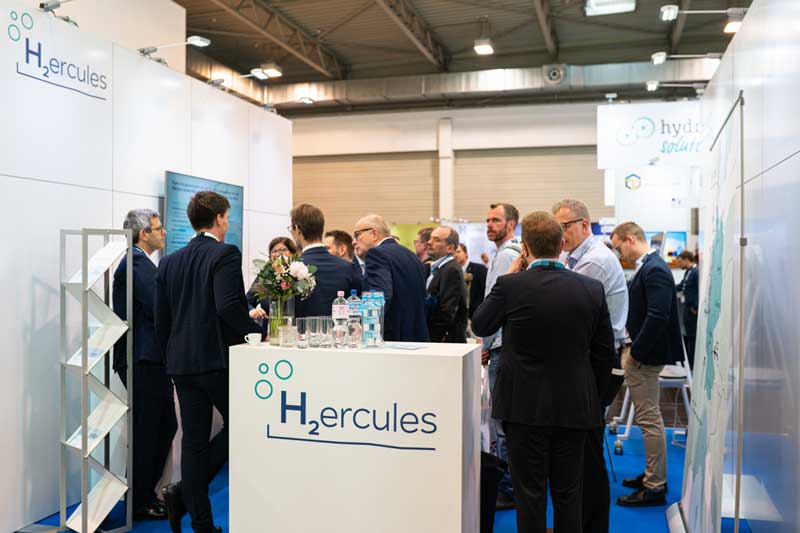 A trade show booth featuring an H2 logo, people engaging in discussions, with water bottles and flowers on display.