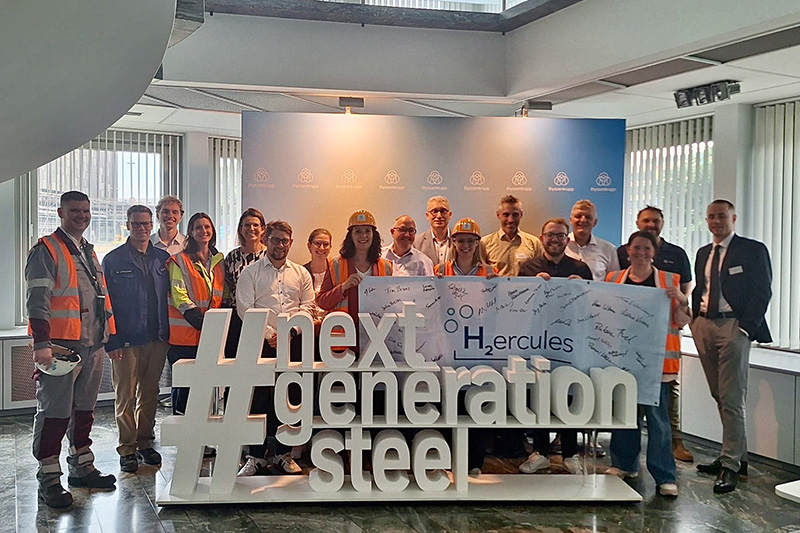 A group of people stand behind a large sculpture reading 'next generation steel' and a banner with 'H2ercules'.