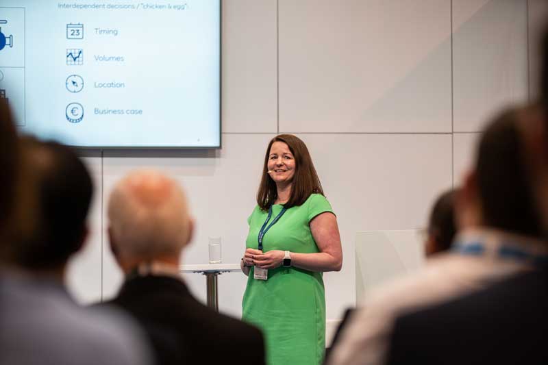 Eine Frau im grünen Kleid präsentiert auf einer Konferenz, mit einem Präsentationsbildschirm, der wichtige Themen im Hintergrund zeigt.