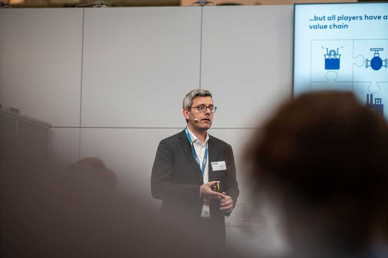 Ein Redner im Anzug präsentiert vor einem Bildschirm, der eine Wertschöpfungskette zeigt, während das Publikum sichtbar ist.