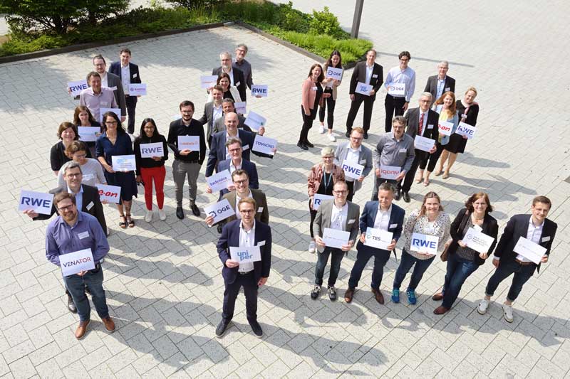 Eine Gruppe von Fachleuten, die Schilder von verschiedenen Unternehmen halten, versammelt sich im Freien an einem sonnigen Tag.