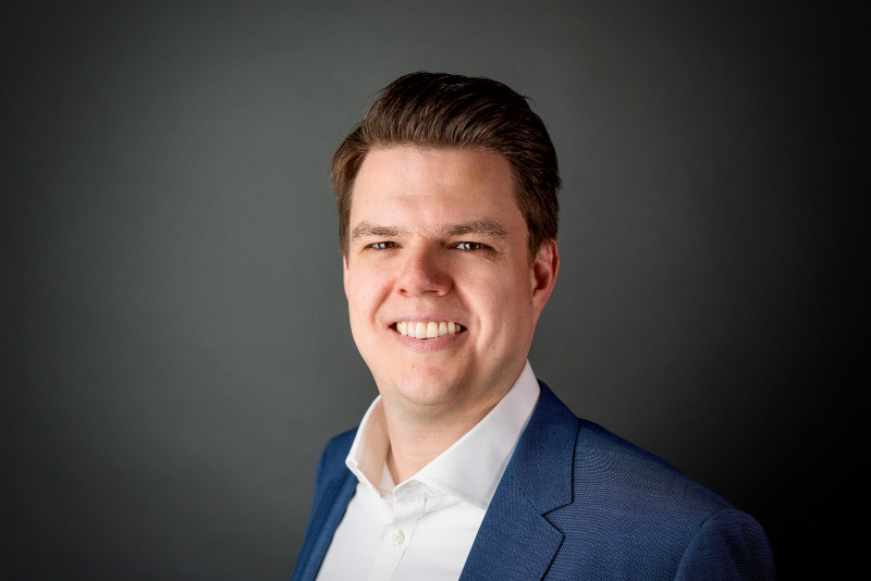 Ein Mann mit kurzem Haar trägt ein blaues Jackett und ein weißes Hemd vor einem dunklen Hintergrund.