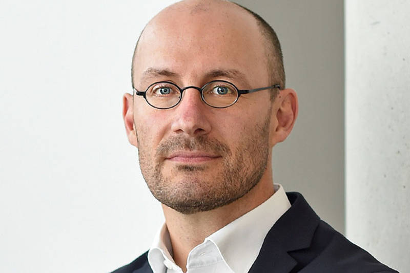 Ein Mann in einem eleganten dunklen Anzug mit einem weißen Hemd steht vor einem hellgrauen Hintergrund.